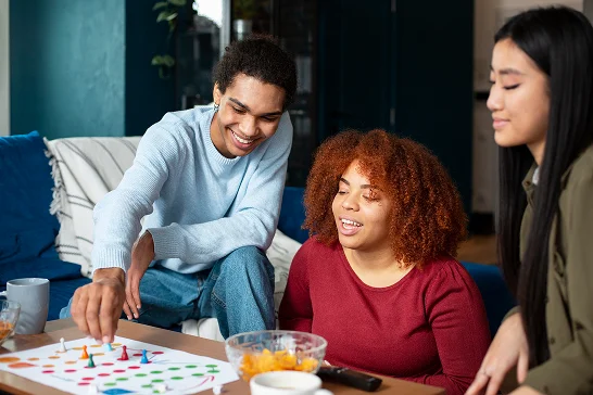 Tre giovani amici sorridenti seduti attorno a un tavolo da gioco mentre giocano a un gioco da tavolo con pedine colorate in un soggiorno accogliente.